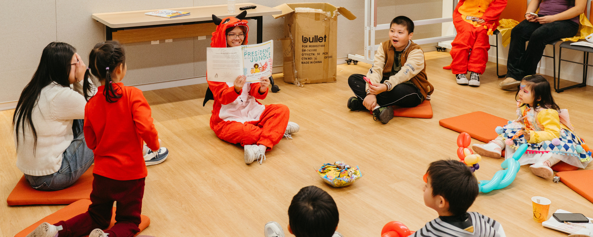 A woman dressed in a red fox costume sits on the floor reading a picture book titled President Junie to a circle of young children, some in costumes, in a brightly lit room with wooden flooring and orange floor cushions.