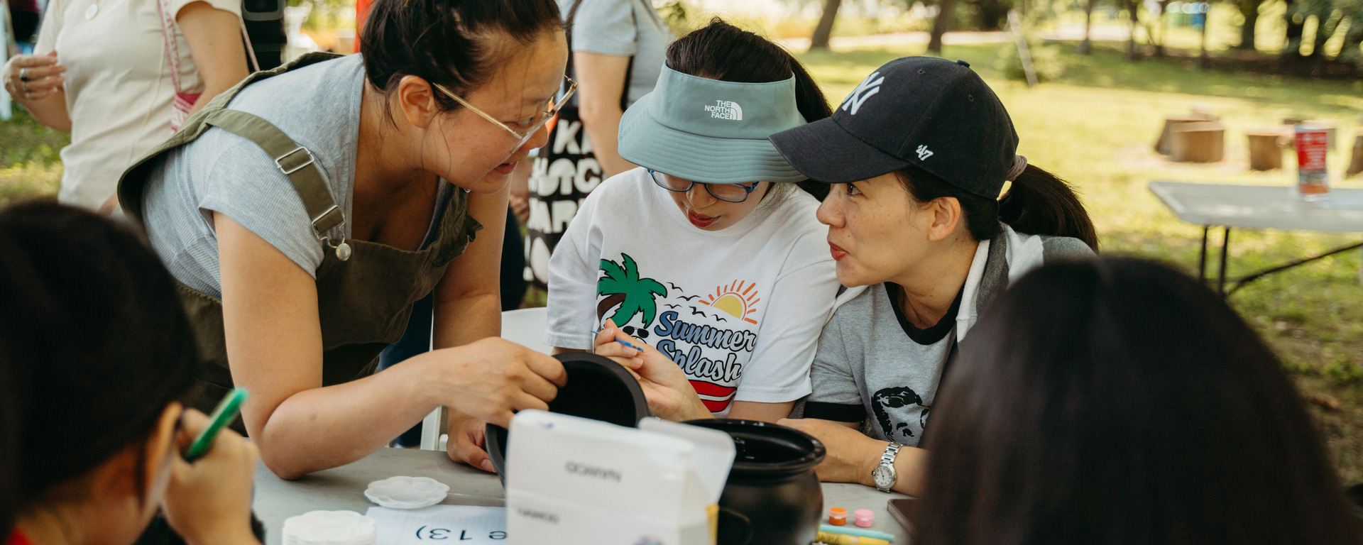 Three women volunteering outdoors at a community event, leaning over a table together with crafts and supplies.