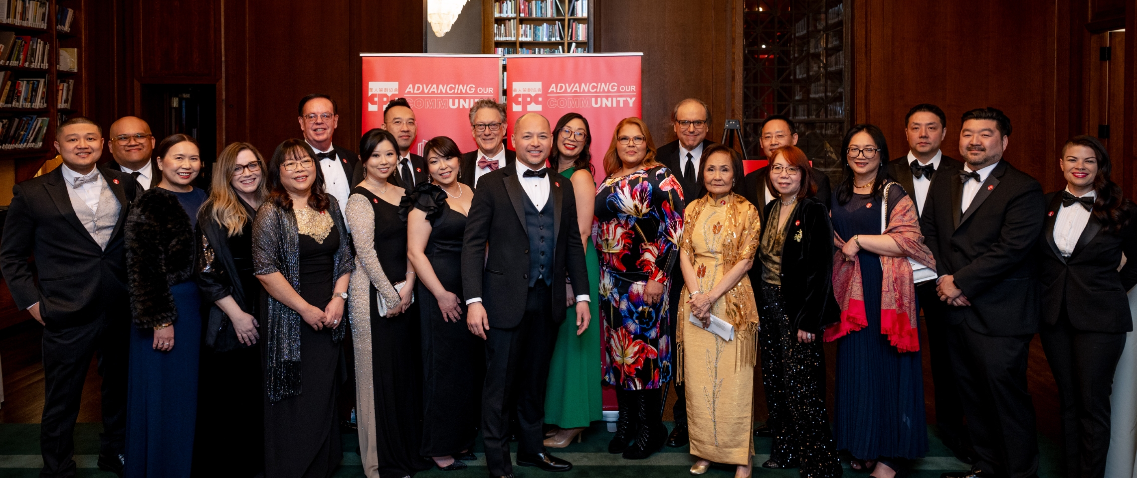 A diverse group of elegantly dressed individuals in formal attire posed together at a gala event for the Chinese-American Planning Council. They stand in front of red banners reading 'ADVANCING OUR COMMUNITY UNITY' in a wood-paneled room with bookshelves
