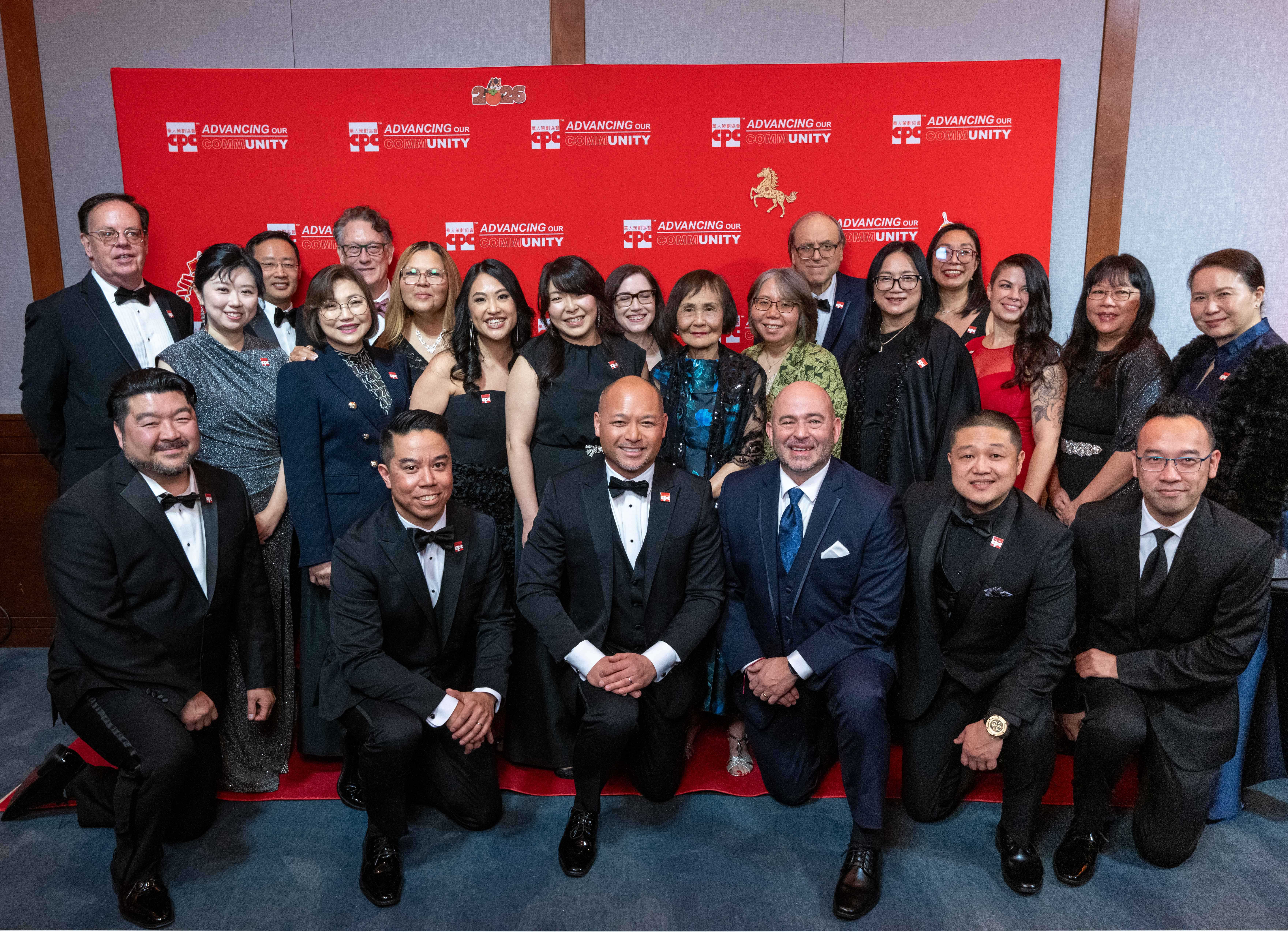 A group of nine well-dressed individuals pose together at a formal event in front of red banners that read "Advancing Our Community." The group includes both men and women wearing suits, tuxedos, and evening gowns, all smiling at the camera
