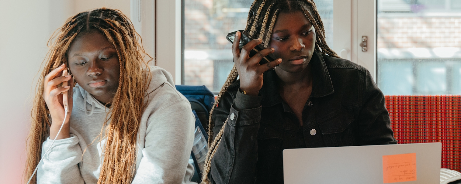 Two teenage girls sit together indoors, each holding a phone to their ear while working; one has a laptop open in front of her.