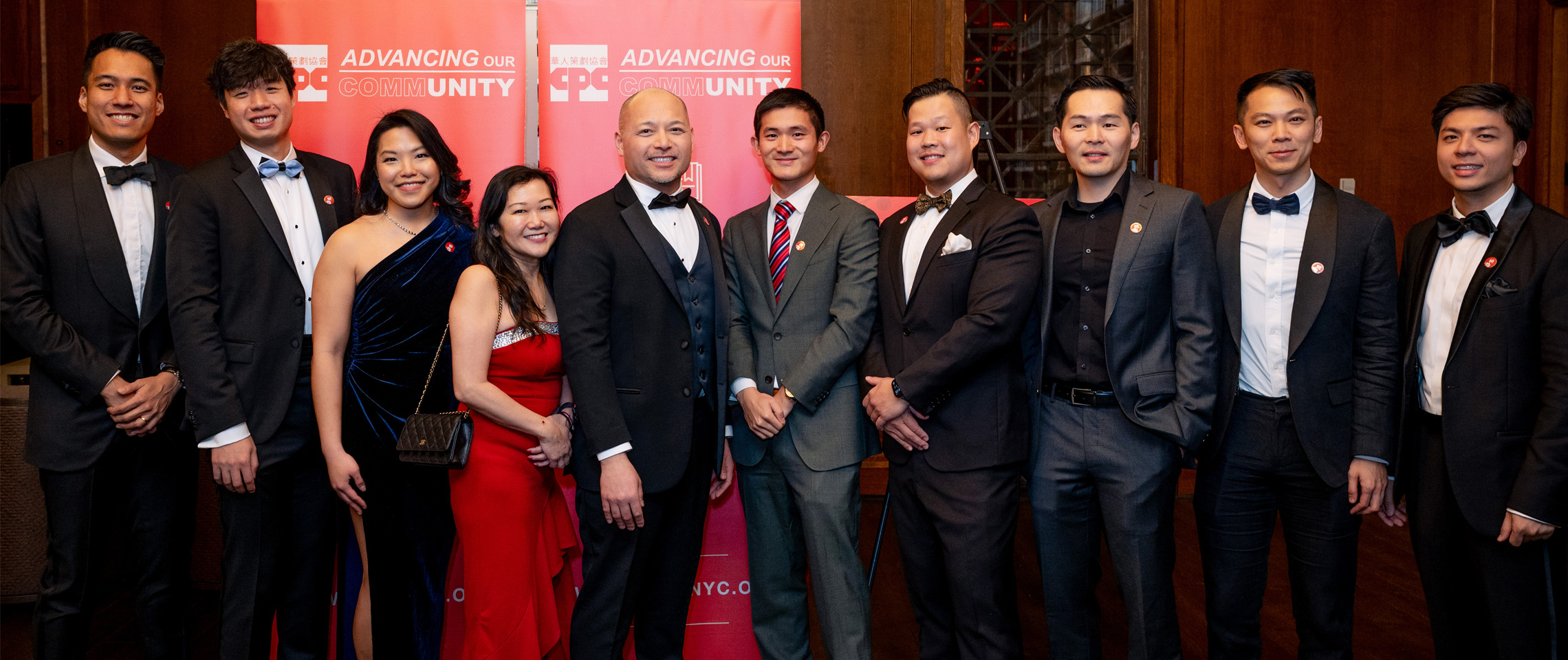 A group of nine well-dressed individuals pose together at a formal event in front of red banners that read "Advancing Our Community." The group includes both men and women wearing suits, tuxedos, and evening gowns, all smiling at the camera